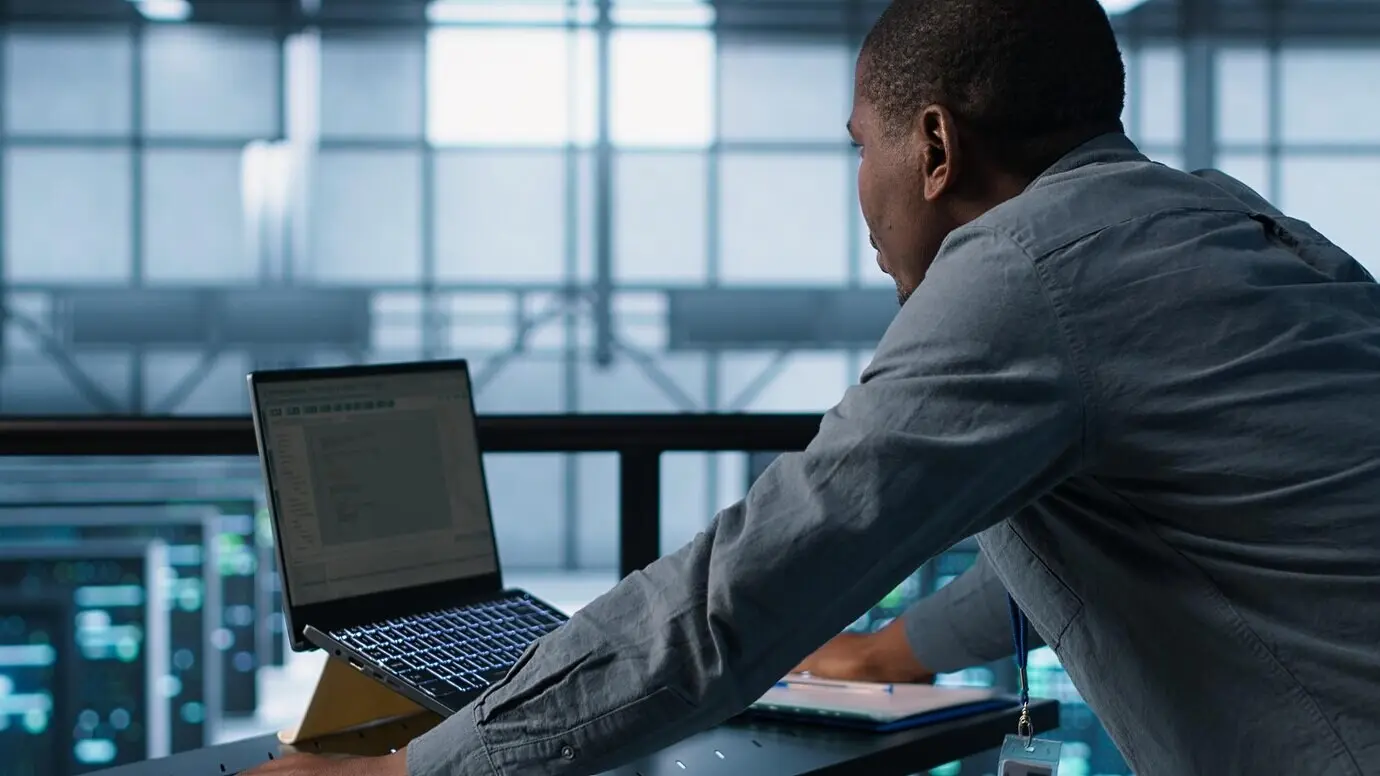 Техник программирует на ноутбуке в центре обработки данных, управляя виртуализацией серверов.